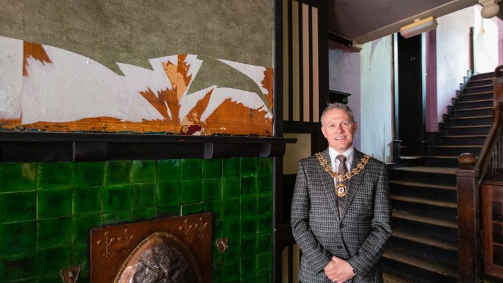 The Town Mayor, wearing his chain of office, stands in 60 King Street where ripped wallpaper can be seen in the foreground