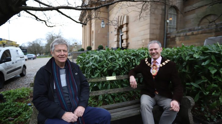 Paul Buttrick and the Town Mayor sit on a bench with a plaque