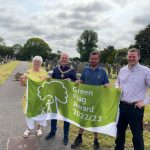Four people, one the town mayor, stand in a cemetery holding up the green flag