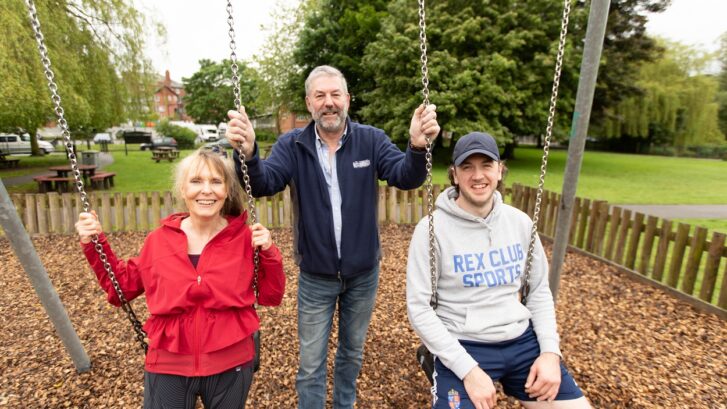 Cllrs Su Russel and Gus Watson sat on swings, with Cllr James McCulloch stood behind holding them