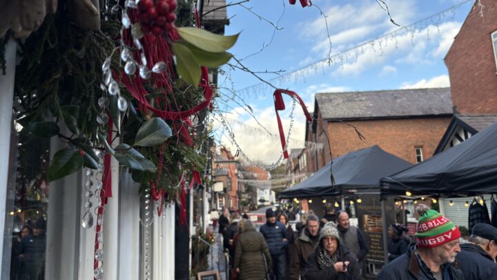 A christmas Wreath hanging from a shop window with christmas market gazebos and festoon lighting and customers perusing the streets.