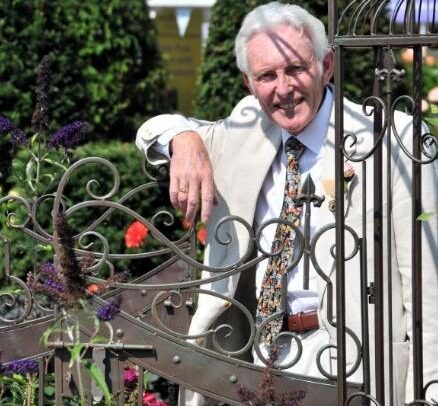 Sam Youd Leaning on garden fence, smiling, dressed in a beige suit and tie.