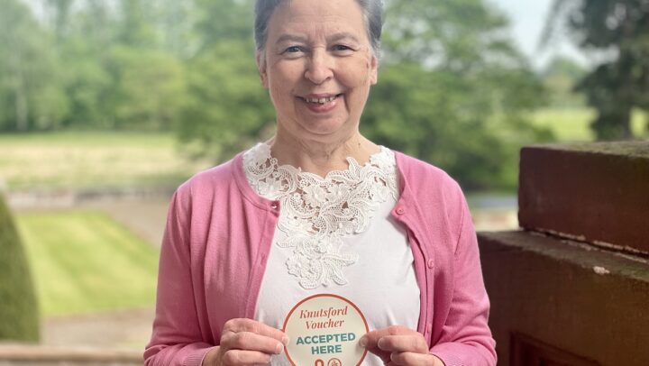 Lady in pink cardigan holding "Knutsford Voucher Accepted Here" sticker
