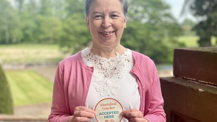 Lady in pink cardigan holding "Knutsford Voucher Accepted Here" sticker